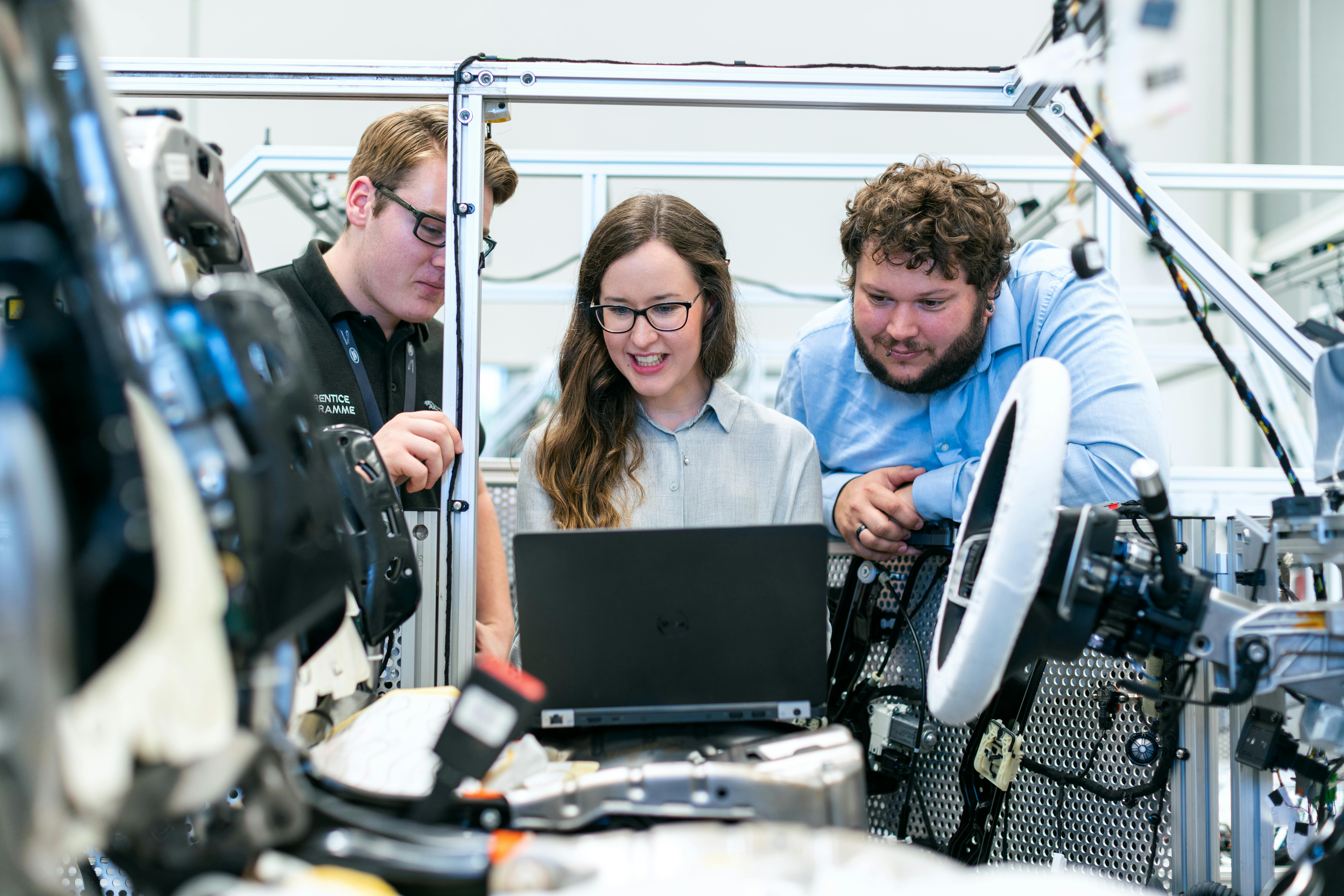 Three engineers working together on a laptop while examining machinery components in a manufacturing lab. Three engineers working together on a laptop while examining machinery components in a manufacturing lab.